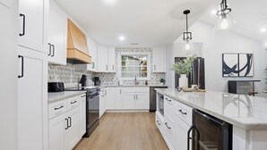 A modern kitchen with white cabinetry, a central island, and a window with blinds. - Shadowlake 4BR House (Charlotte)