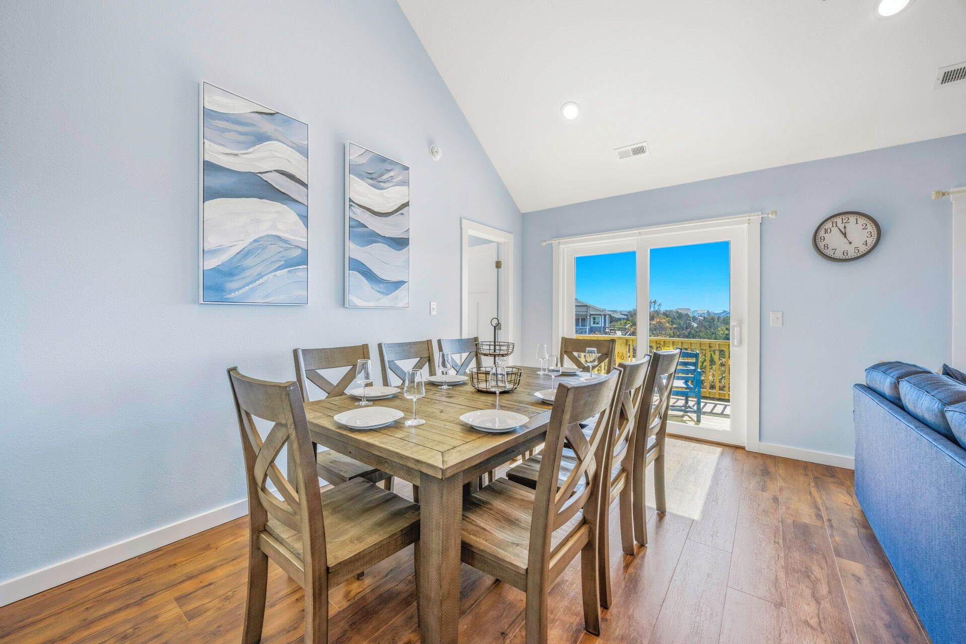 A dining area with a wooden table and chairs, a view through a glass door, and a clock on the wall.