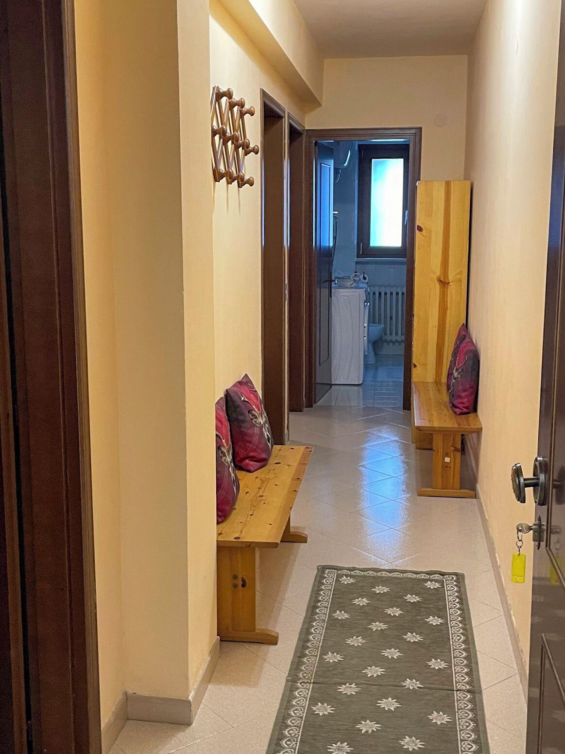 Un couloir avec des bancs en bois et un tapis, qui mène à une pièce dotée d’une fenêtre et d’un radiateur.