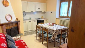 A cozy living room with a fireplace, a dining area with a table and chairs, and a kitchen with cabinets and a window. - Casa Vacanze - Villa Stefania (Pescocostanzo)