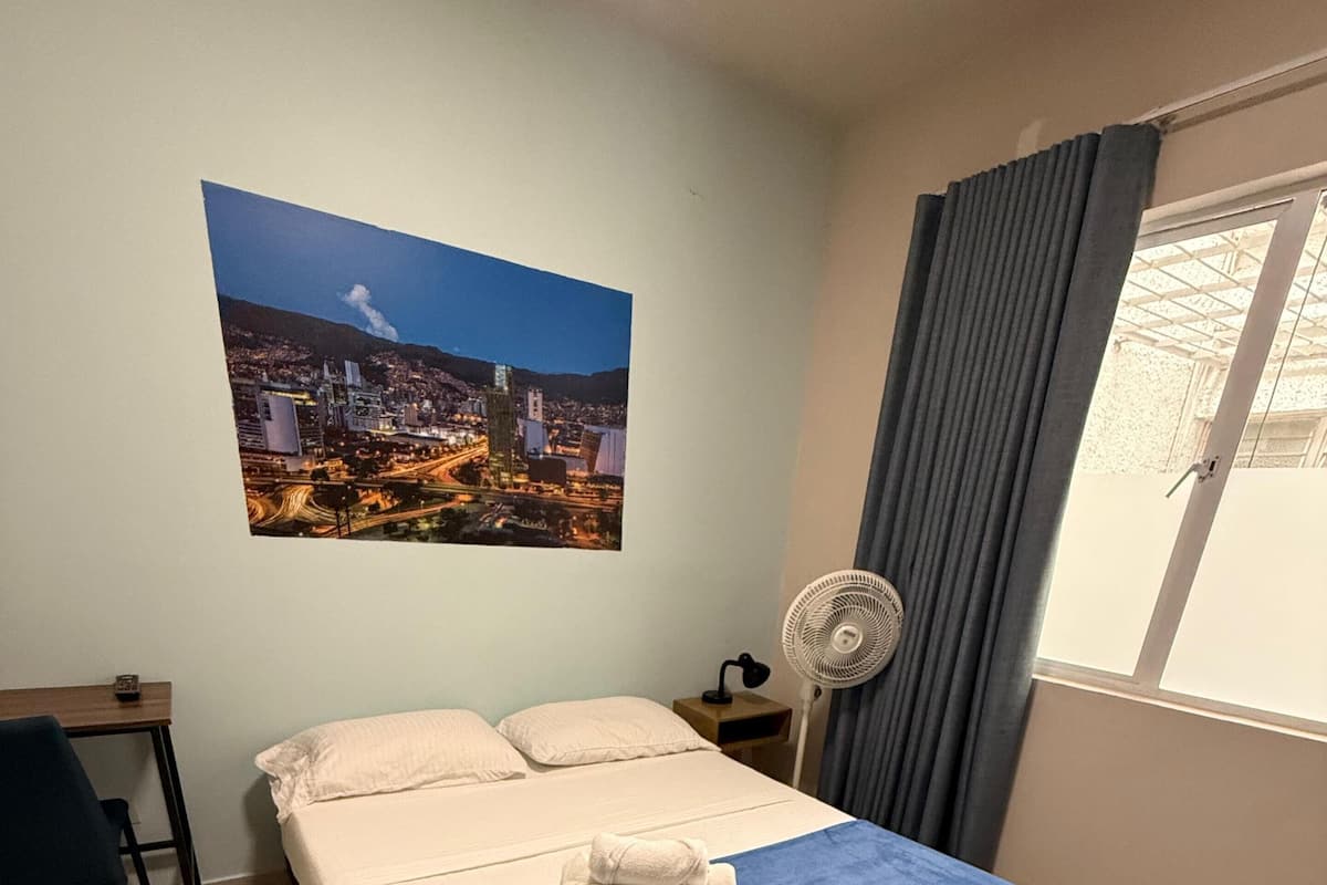 A bedroom with a bed, a desk, a chair, a nightstand, a wall-mounted fan, and a window with curtains.