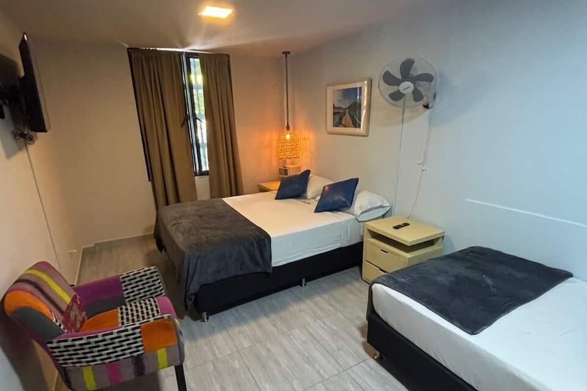 A compact hotel room with two beds, a sofa, a TV, a fan, and a window with curtains.