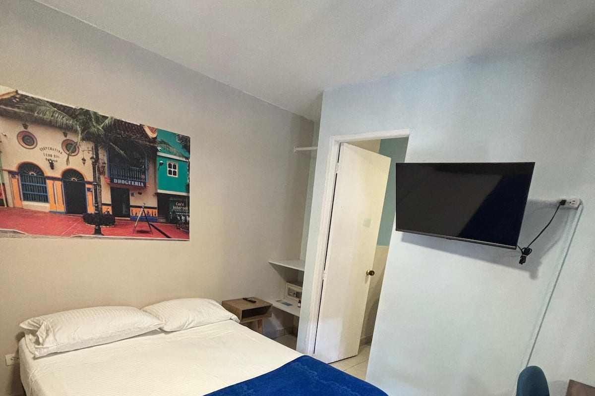 A room with a bed, a blue blanket, a chair, a TV mounted on the wall, and a painting of a building.
