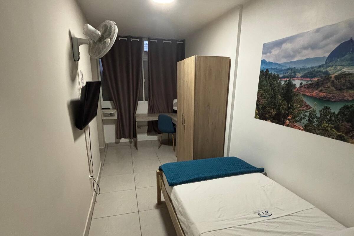 A small, tidy room with a bed, a desk, a wardrobe, and a fan.