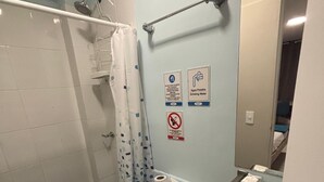 Un baño con inodoro, lavamanos, ducha y espejo. Se observan letreros en la pared que indican que no hay agua caliente y que hay agua potable disponible.