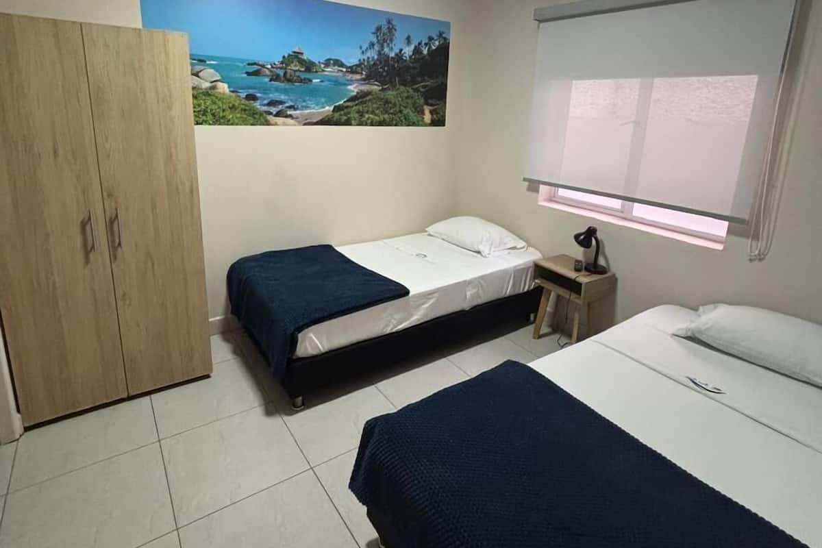 A bedroom with two beds, a wardrobe, a painting of a beach scene, a ceiling fan, and a window with blinds.