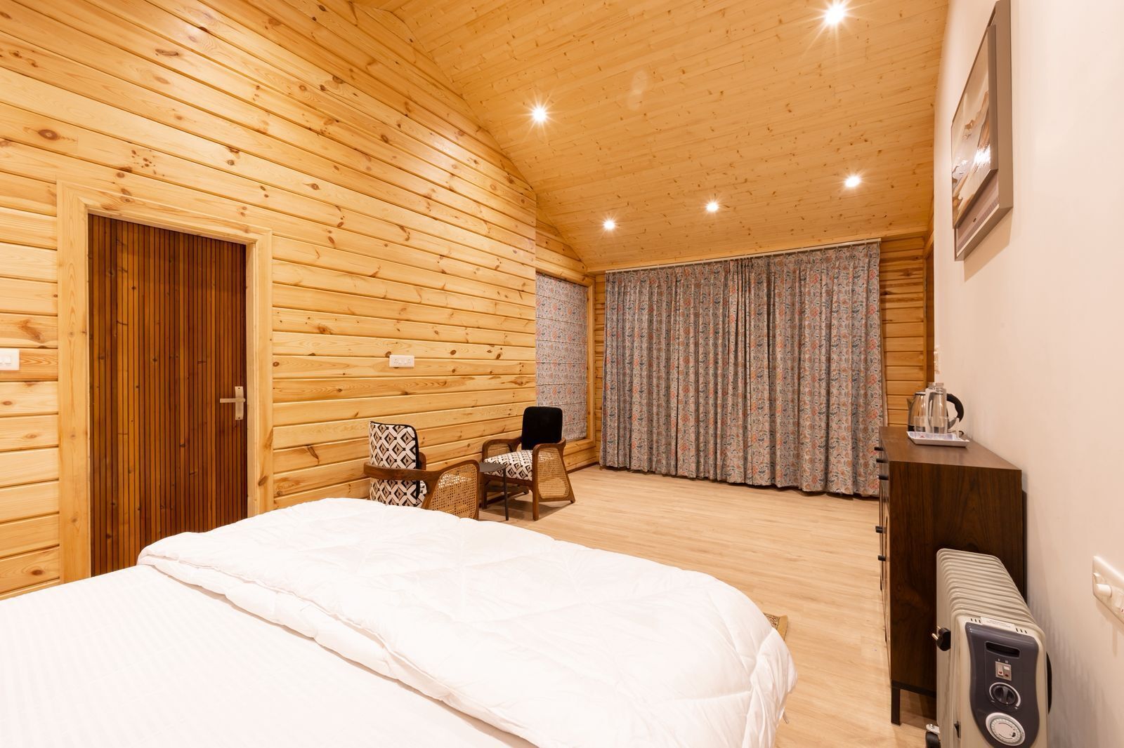 Habitación tipo cabaña de madera con cama, dos sillas, una mesa con hervidor y un ventanal grande con cortinas.