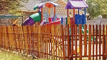 Una valla de madera frente a un parque infantil con un tobogán y estructuras para escalar.