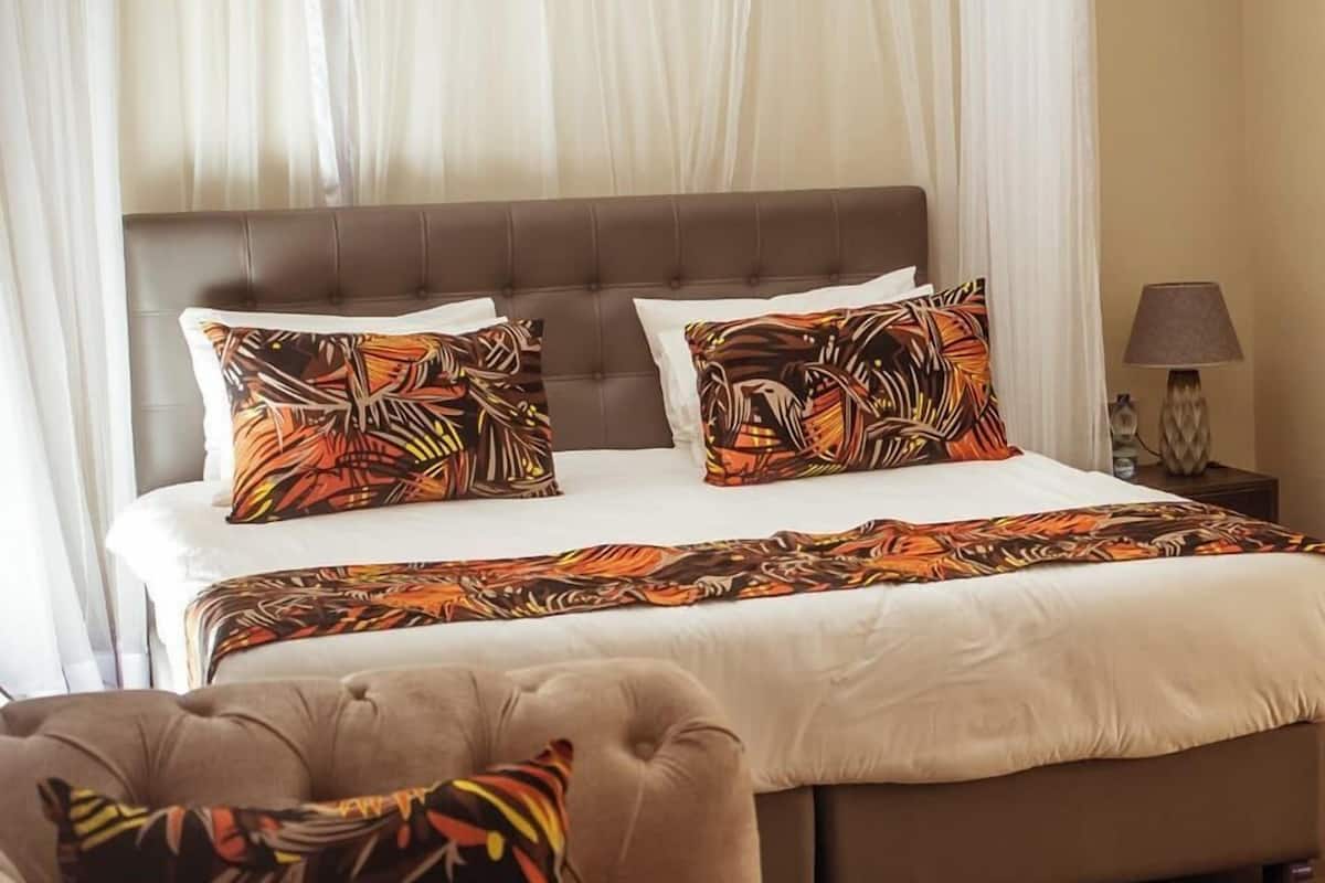 A bedroom with a large bed, two pillows with a tropical pattern, a brown headboard, and a bedside table with a lamp.