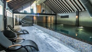 A modern indoor swimming pool with a tiled edge, a sloped ceiling with wooden beams, and lounge chairs along the poolside. - Upper Home Farm (Church Stretton)