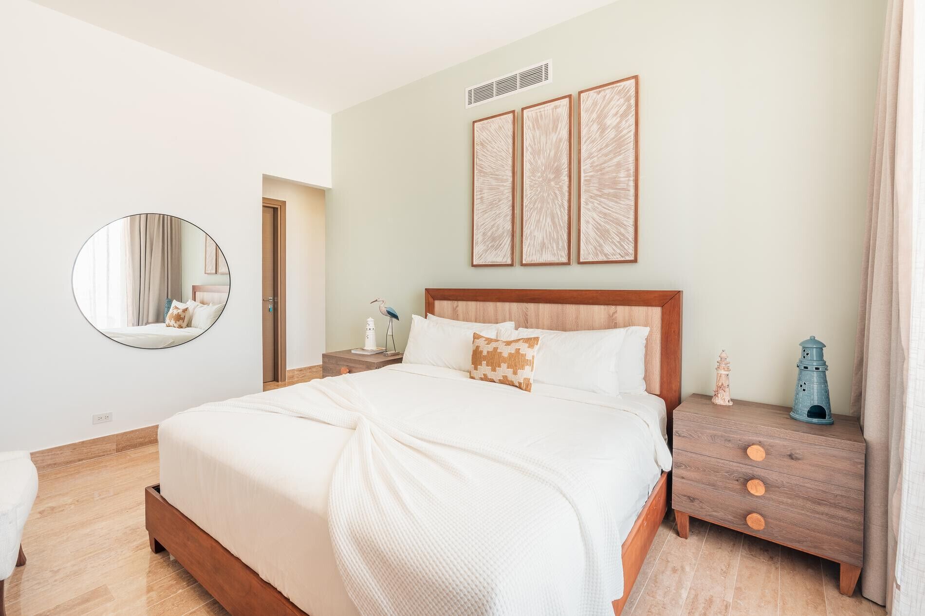 A hotel room with a large bed, a round mirror, a nightstand with decorative items, and a wall with framed artwork.