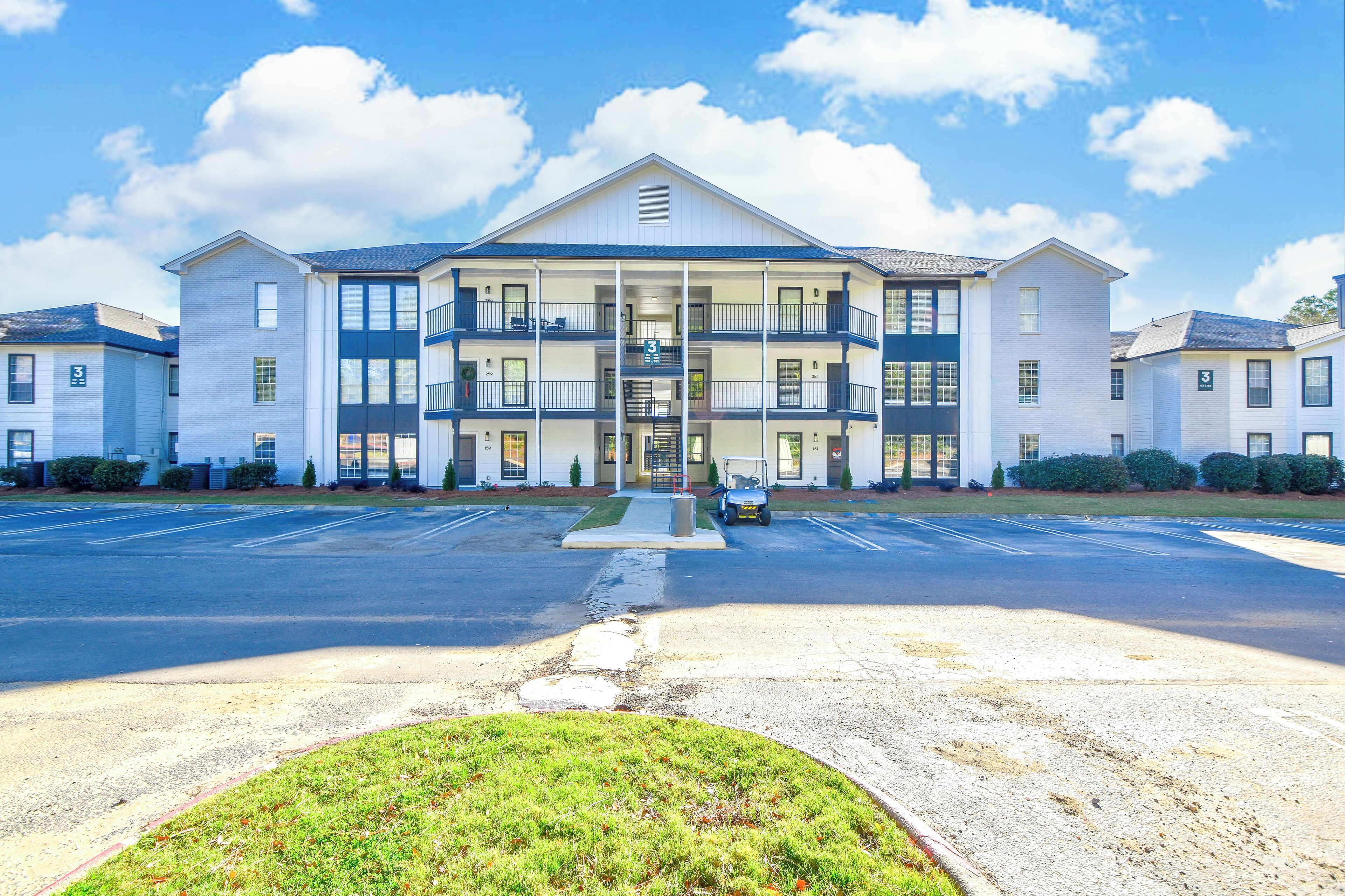 A multi-story building with a central entrance, balconies, and a parking lot in front.
