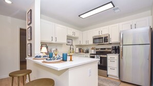 A modern kitchen with white cabinets, a stainless steel refrigerator, a microwave, and an oven. There are two stools at the island and a small rug on the floor. - New! Shug's Station, 2 BR, 2BA condo close to Auburn University, sleeps 4! (Auburn)