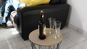 Un hombre sentado en un sofá negro leyendo un periódico, con una botella y vasos sobre una mesa redonda frente a él.