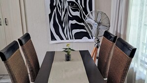 A dining table with a beige tablecloth, surrounded by wicker chairs with black and brown cushions. A large zebra print artwork is on the wall above the table. - Cozy, quiet 1-bedroom pet-friendly cottage in the equestrian heart of the metro. (Midrand)