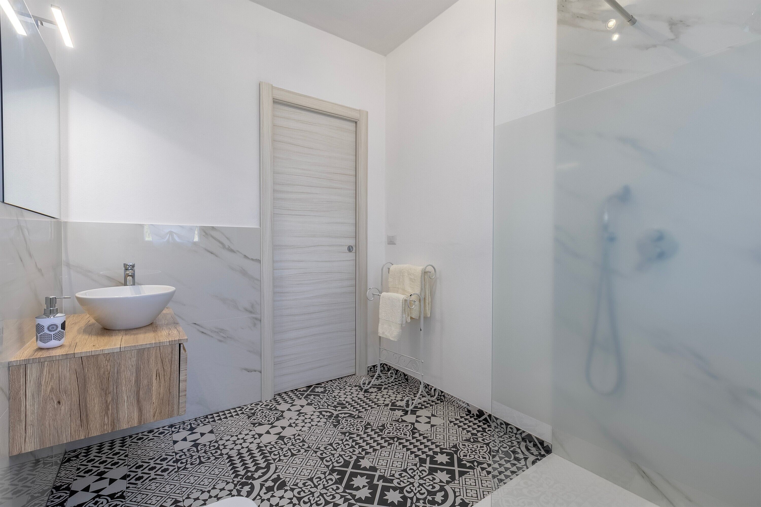 Ein Badezimmer mit einem weißen Waschbecken, einem Holztischchen, einem Musterboden und einer Duschkabine aus Glas.