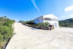 Un edificio moderno blanco con techo curvo, entrada pavimentada y zona ajardinada con rocas y arbustos.