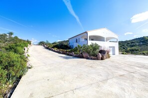 Edifici modern de color blanc amb un teulat corbat, una calçada asfaltada i una zona enjardinada amb roques i arbustos.