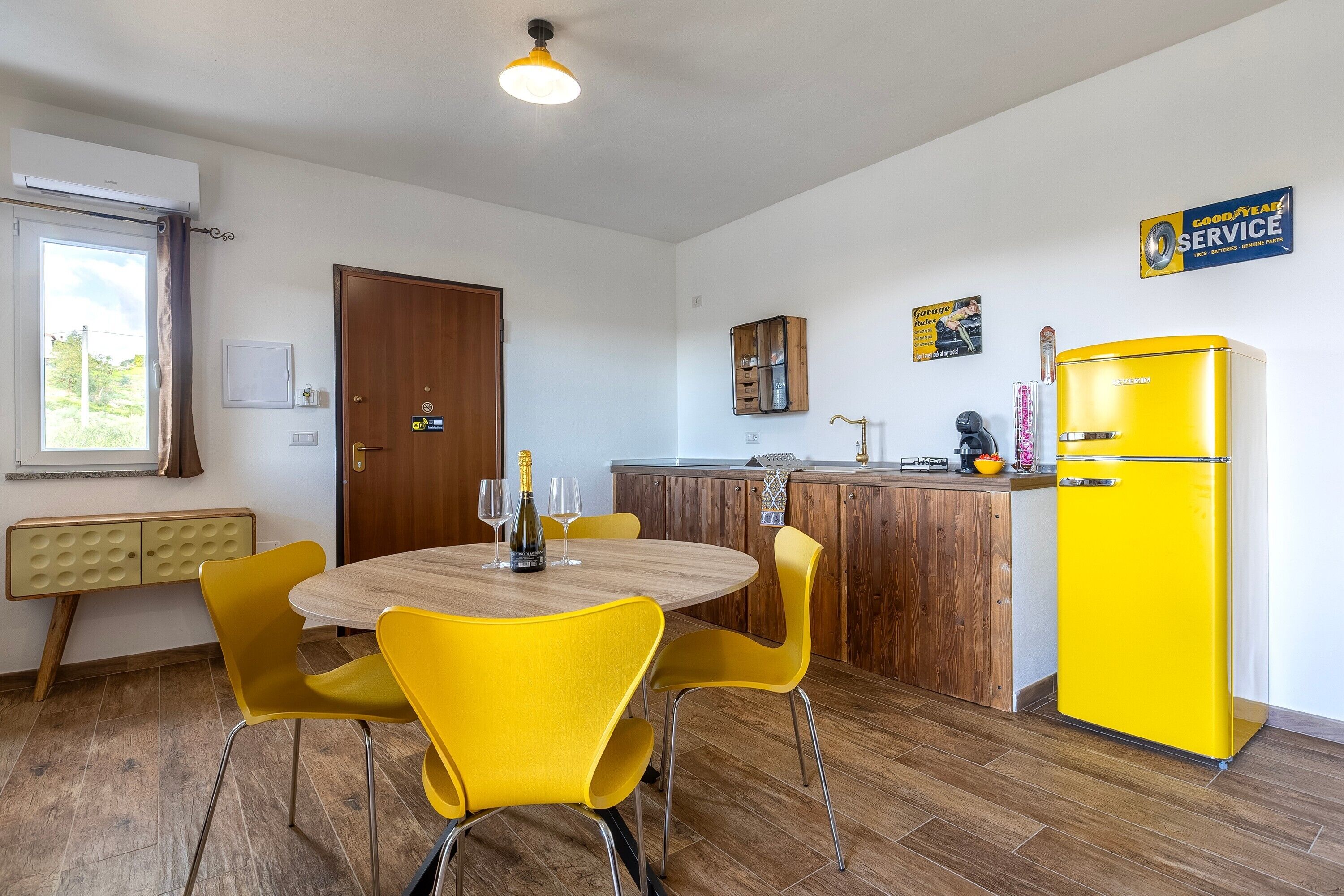 A kitchen with a yellow refrigerator, wooden cabinets, a round dining table with yellow chairs, and a window with curtains.