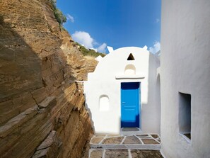 A narrow pathway leading to a white building with a blue door, set against a rocky hillside under a clear sky.