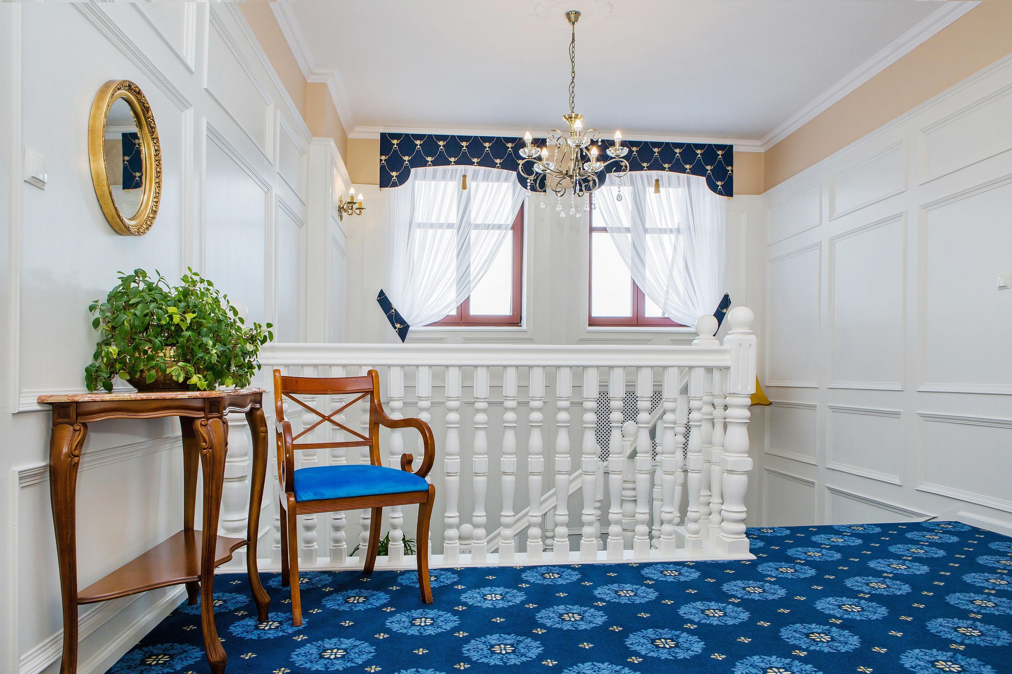 Une pièce avec un tapis bleu à motifs, une chaise en bois avec un coussin bleu, une table en bois avec une plante, une rambarde blanche et un lustre.
