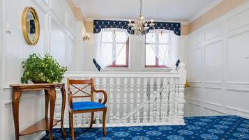 Une pièce avec un tapis bleu à motifs, une chaise en bois avec un coussin bleu, une table en bois avec une plante, une rambarde blanche et un lustre.