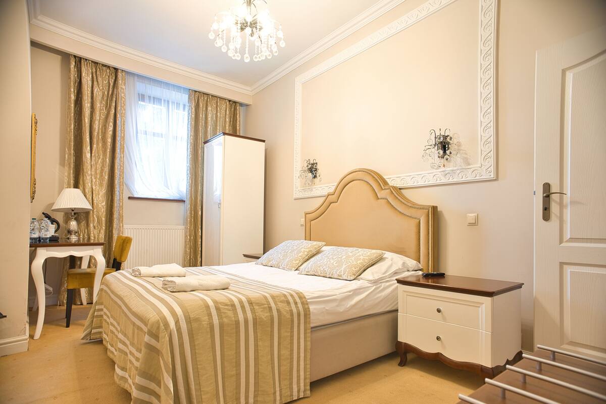 A well-lit bedroom with a large bed, a bedside table, a desk with a lamp, and a chair.