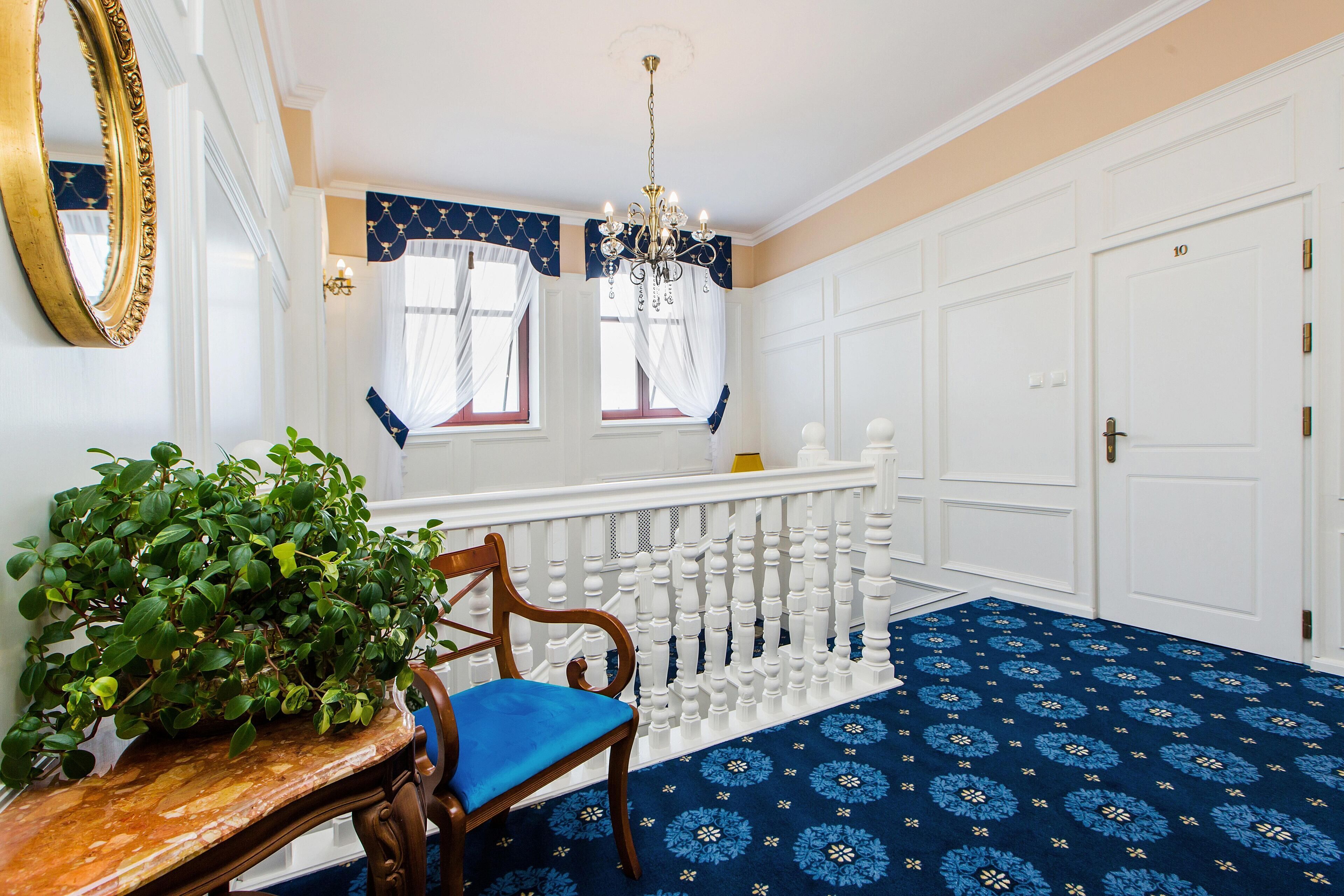 Un couloir avec un tapis à motifs bleus, une chaise en bois avec un coussin bleu, une plante en pot, une rambarde blanche et un lustre.