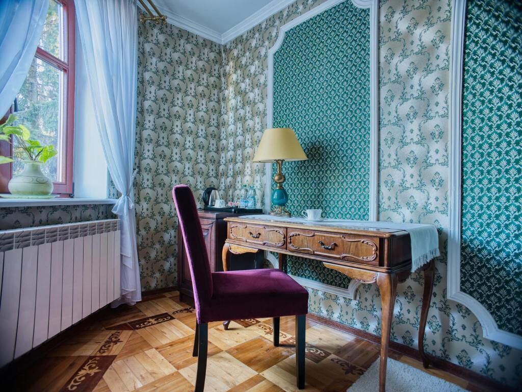 A room with patterned wallpaper, a wooden desk, a chair, a lamp, and a window with curtains.