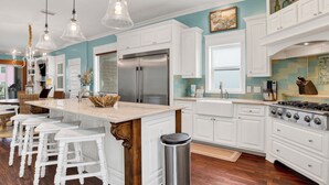A modern kitchen with white cabinetry, a stainless steel refrigerator, a gas stove, and a large island with bar stools. - Gulf Views | Private Pool | Outdoor Kitchen (Fort Morgan)