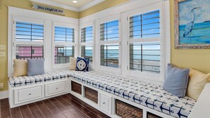 A bay window seating area with multiple windows, a bench with nautical-themed cushions, and a painting of a bird on the wall. - Gulf Views | Private Pool | Outdoor Kitchen (Fort Morgan)