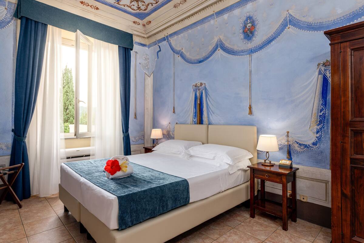 Una habitación de hotel con una cama grande, dos mesitas de noche, una ventana con cortinas y un mural decorativo en la pared.