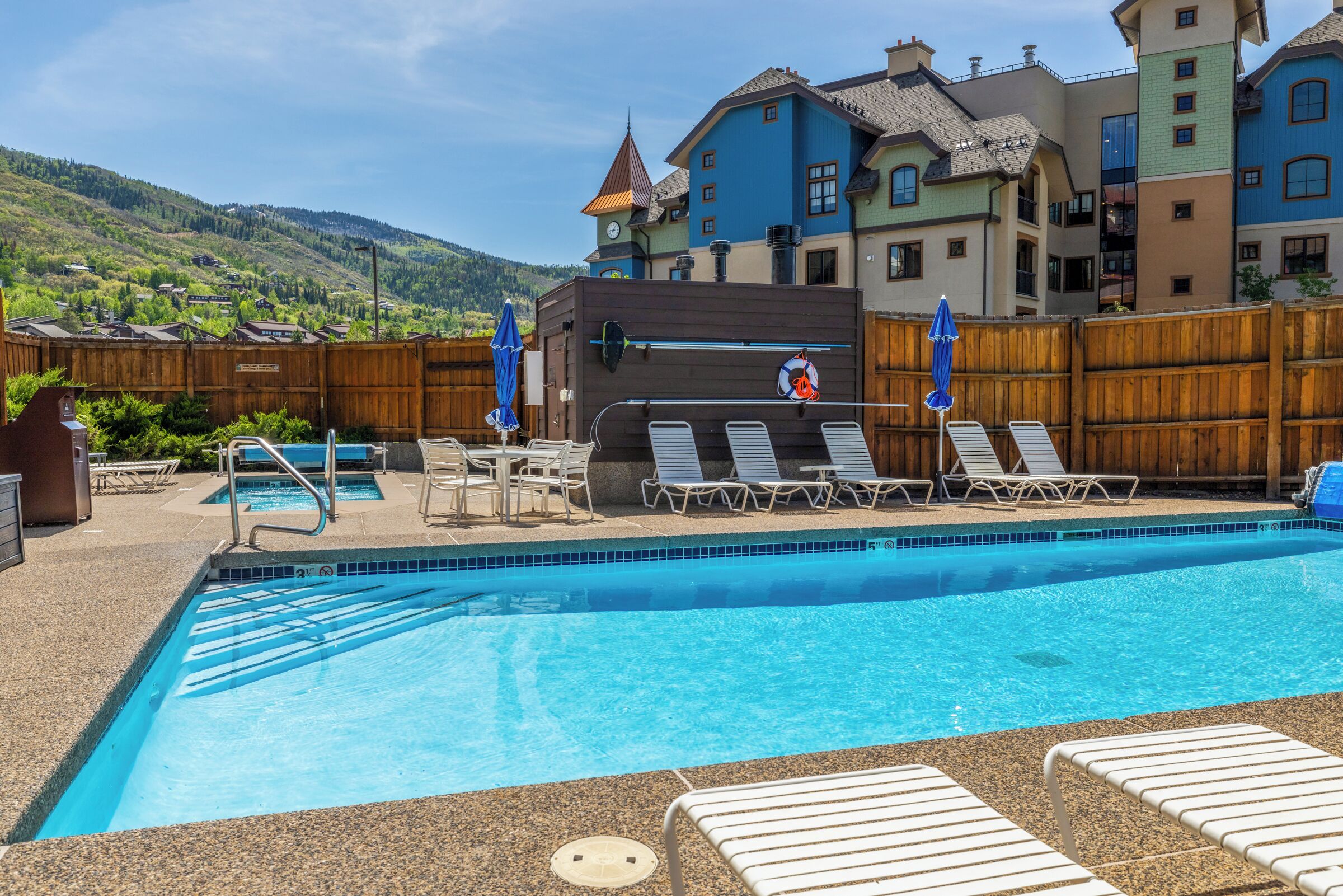 A swimming pool with lounge chairs and umbrellas, surrounded by a wooden fence.