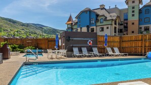 A swimming pool with lounge chairs and umbrellas, surrounded by a wooden fence. - Base Area Condo | Steps to Ski | Pool and Hot Tub (Steamboat Springs)
