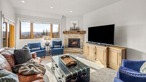 A living room with a fireplace, a large flat-screen TV, leather sofas, and a view of the outdoors. - Base Area Condo | Steps to Ski | Pool and Hot Tub (Steamboat Springs)