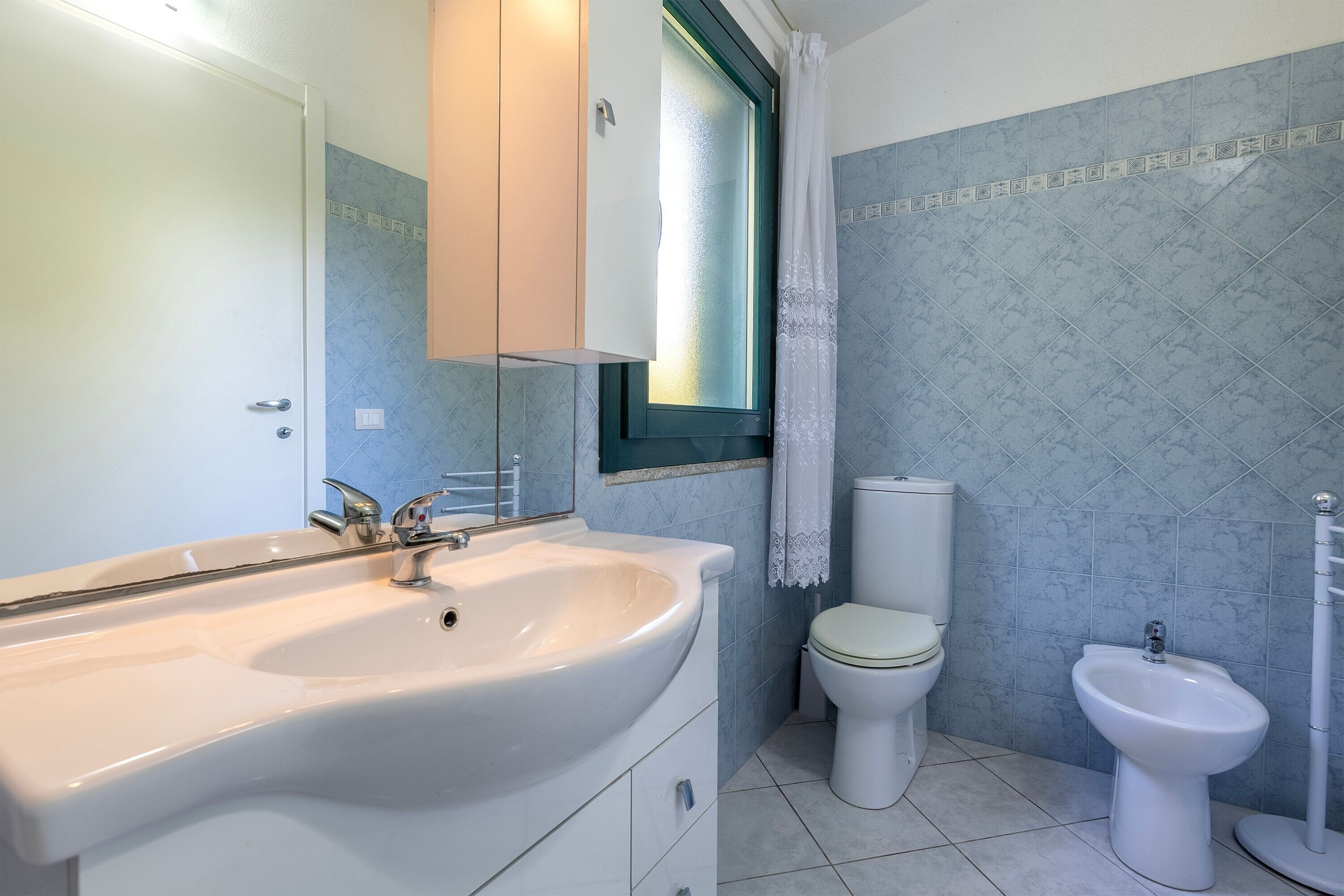 A bathroom with a sink, toilet, bidet, and a window with a curtain.