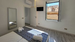A modern hotel room with a bed, a mirror, a window with a view of buildings, and an air conditioning unit. - Sara luxury apartment 3 (Sorrento)