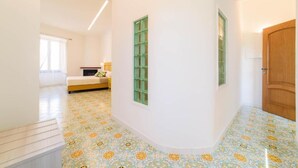 A hallway with patterned floor tiles, a bed with a green window, and a wooden door. - Sara Luxury Rooms - Positano Room (Meta)
