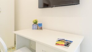 A white desk with a flat-screen TV mounted above it, a small potted plant, and a couple of books on the desk. - Sara Luxury Rooms - Positano Room (Meta)