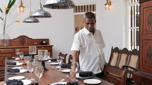 Un comedor con una mesa de madera dispuesta para una comida, incluyendo platos, vasos y cubiertos. Hay lámparas colgantes sobre la mesa, y se ve un armario de madera con un panel decorativo al fondo.