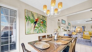 A dining area with a wooden table, woven placemats, and a large painting of parrots. - Wavy Seas - Tennis Villa 77A (Miramar Beach)