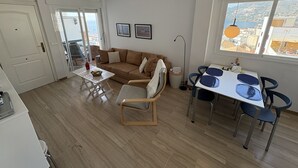 Ein Wohnzimmer mit einer Couch, einem Esstisch mit Stühlen, einem kleinen Tisch mit einer Flasche und Obst sowie Blick auf die Stadt durch große Fenster.