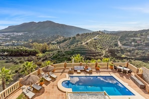 A pool with lounge chairs and umbrellas, surrounded by a stone wall and overlooking a hilly landscape.