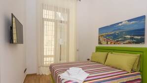 Een slaapkamer met een groen hoofdeinde, een kleurrijk gestreept dekbed en een schilderij van een kustplaats op de muur.