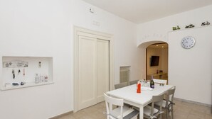 Een eetgedeelte met een witte tafel en stoelen, een klok aan de muur, en een deur die leidt naar een andere kamer.