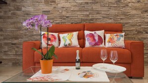 Een moderne woonkamer met een oranje bank, decoratieve kussens, een glazen salontafel, een potplant en twee wijnglazen.