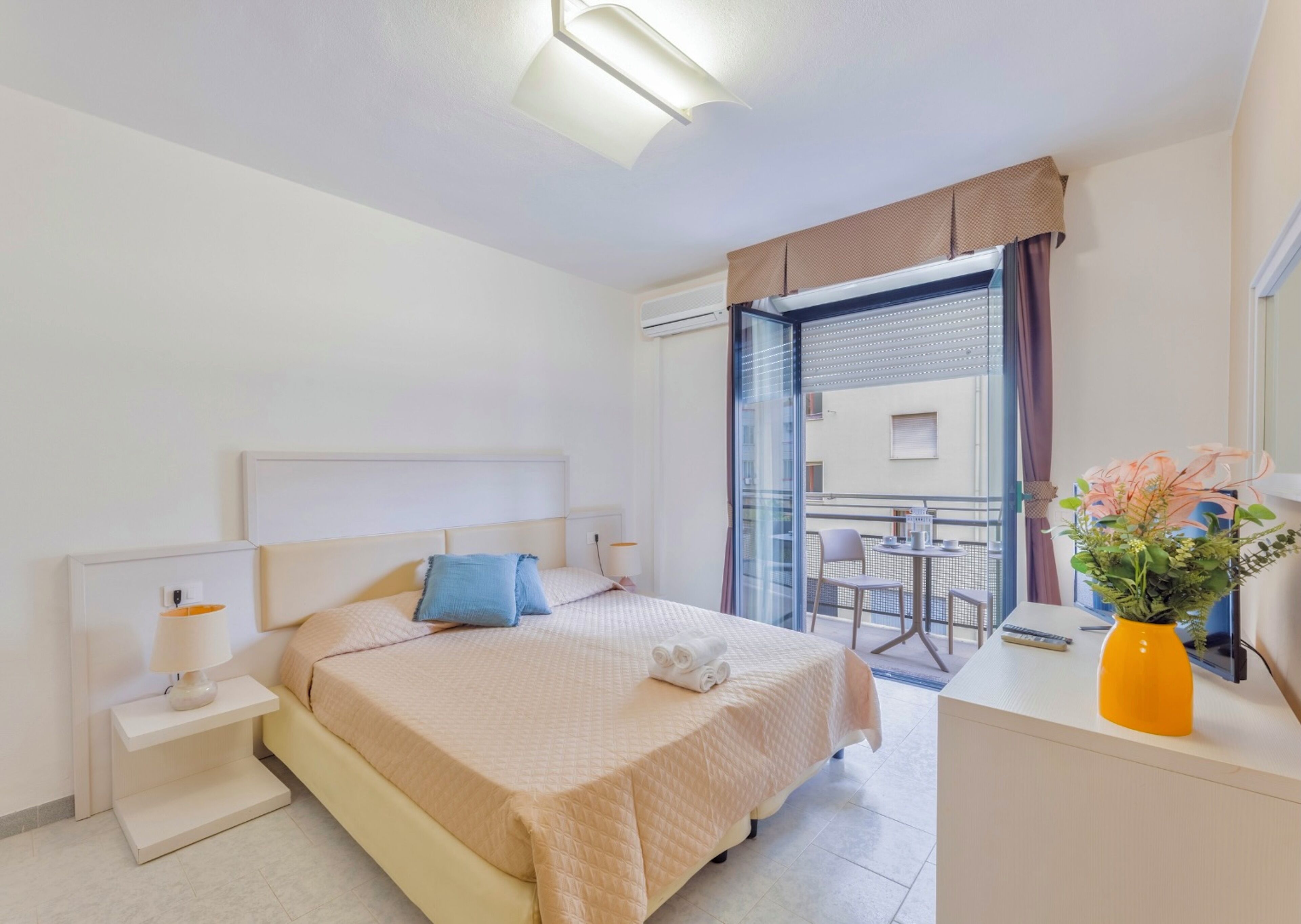 A hotel room with a bed, bedside table, lamp, and a small balcony with a table and chairs.