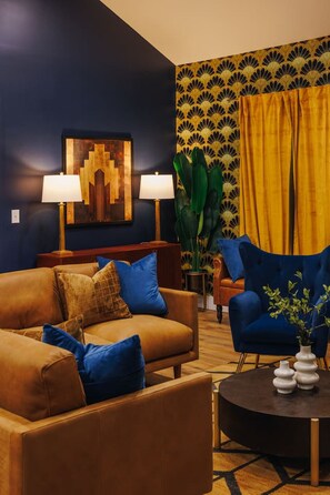 Sala de estar com sofá marrom, poltrona azul e mesa de centro redonda. Há duas luminárias, uma planta grande e uma parede com cortina amarela.
