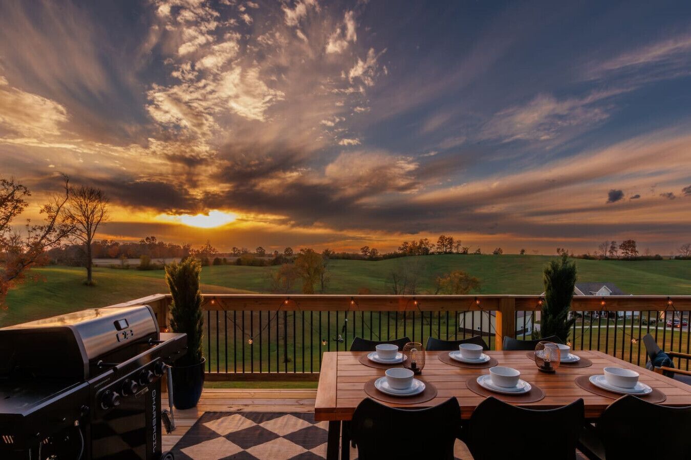 Eine Terrasse mit Grill und Esstisch für sechs Personen, mit Blick auf ein Wiesenfeld und einen Sonnenuntergang.