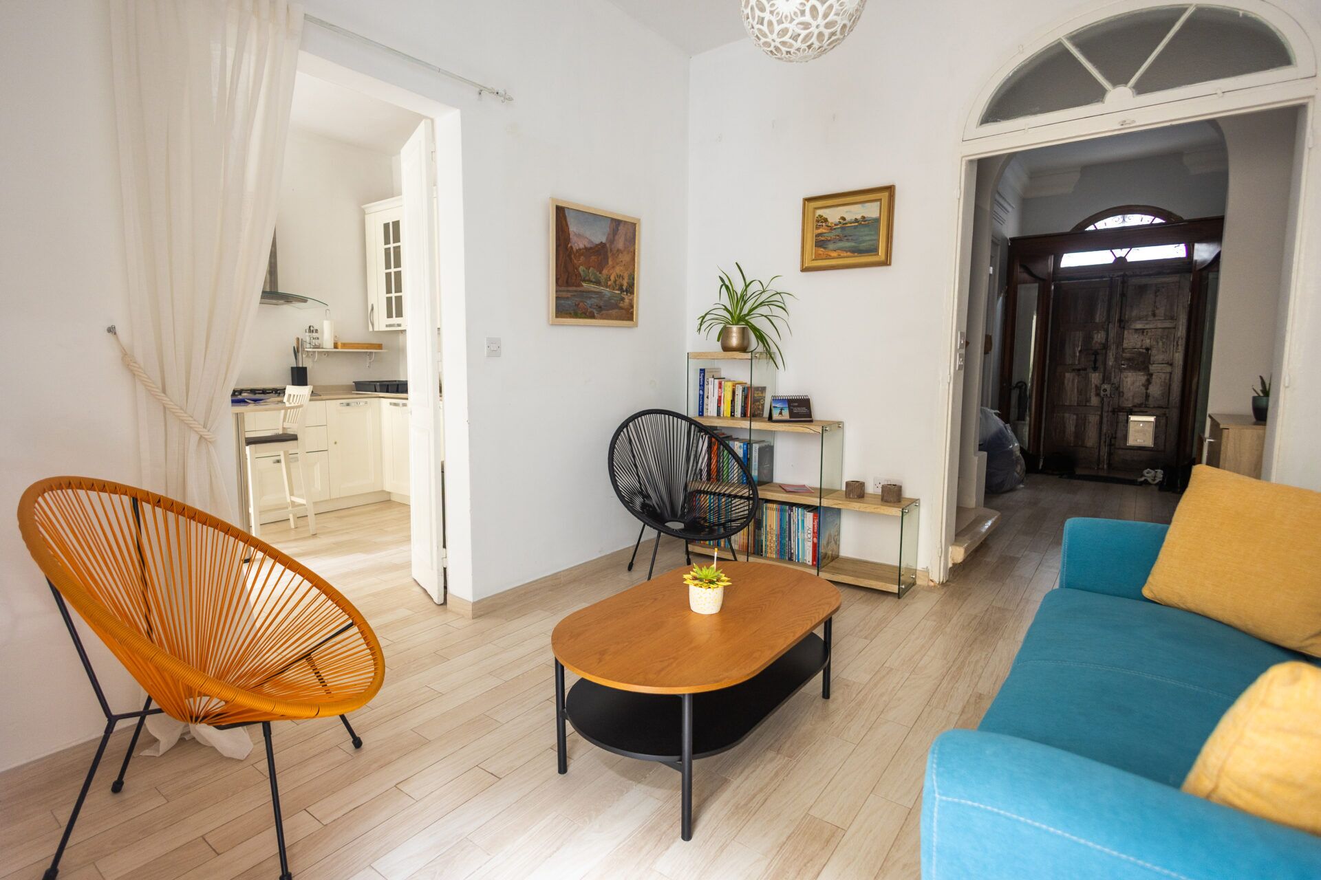 Een woonkamer met een houten salontafel, een blauwe bank, een rieten fauteuil en een boekenkast met boeken en een potplant.